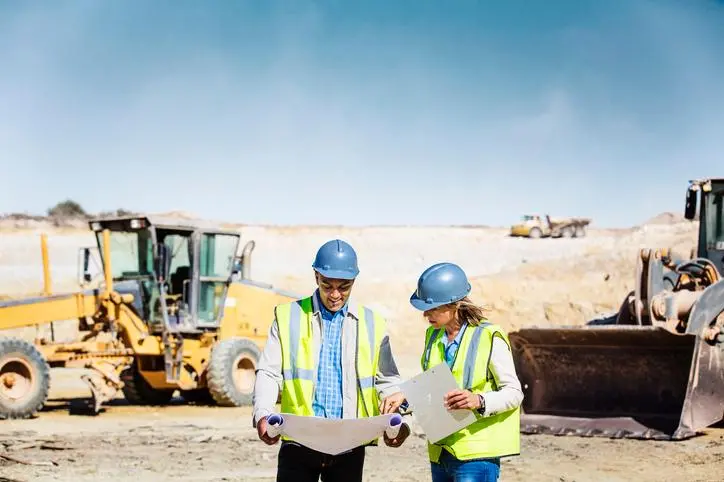 Male and female architects examining blueprint at quarry with bulldozers in background Source: Zawya.com